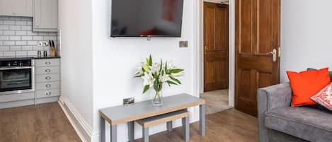 Een moderne woonkamer met een grijze bank, een flatscreen-televisie aan de muur, een houten deur en een keuken met grijze kastjes en een inbouwoven.
