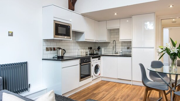 A modern kitchen with white cabinetry, a built-in microwave, a kettle, and a washing machine. There is a dining area with a glass table and chairs. - The Tunstall (Durham)