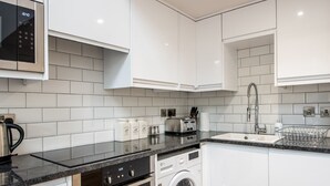 A modern kitchen with white cabinets, a stainless steel microwave, a built-in oven, a washing machine, and a sink with a dish rack. - The Tunstall (Durham)