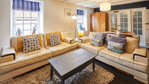 A living room with a beige sectional sofa, a coffee table, and a chandelier. There is a window with a view of people outside. - Host Stay Dotty s Coastal Retreat (Saltburn-by-the-Sea)