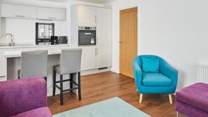 A modern kitchen with white cabinets, a microwave, and an oven. There is a wooden door and a blue armchair in the living area. - Host Stay The Globe Apartment 3 (Guisborough)