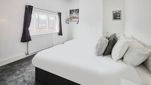 A bedroom with a large bed, a window with curtains, a radiator, a wall-mounted shelf with a picture, and a framed picture on the wall. - Host Stay Church Street (Warrington)