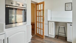 A compact kitchen with white cabinetry, a built-in oven, a wooden-framed door, a white countertop, and a stool. - Host Stay Church Street (Warrington)