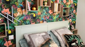 Une chambre à coucher avec un papier peint aux motifs tropicaux éclatants, une tête de lit bleu clair et de nombreuses étagères ornées d’objets décoratifs.