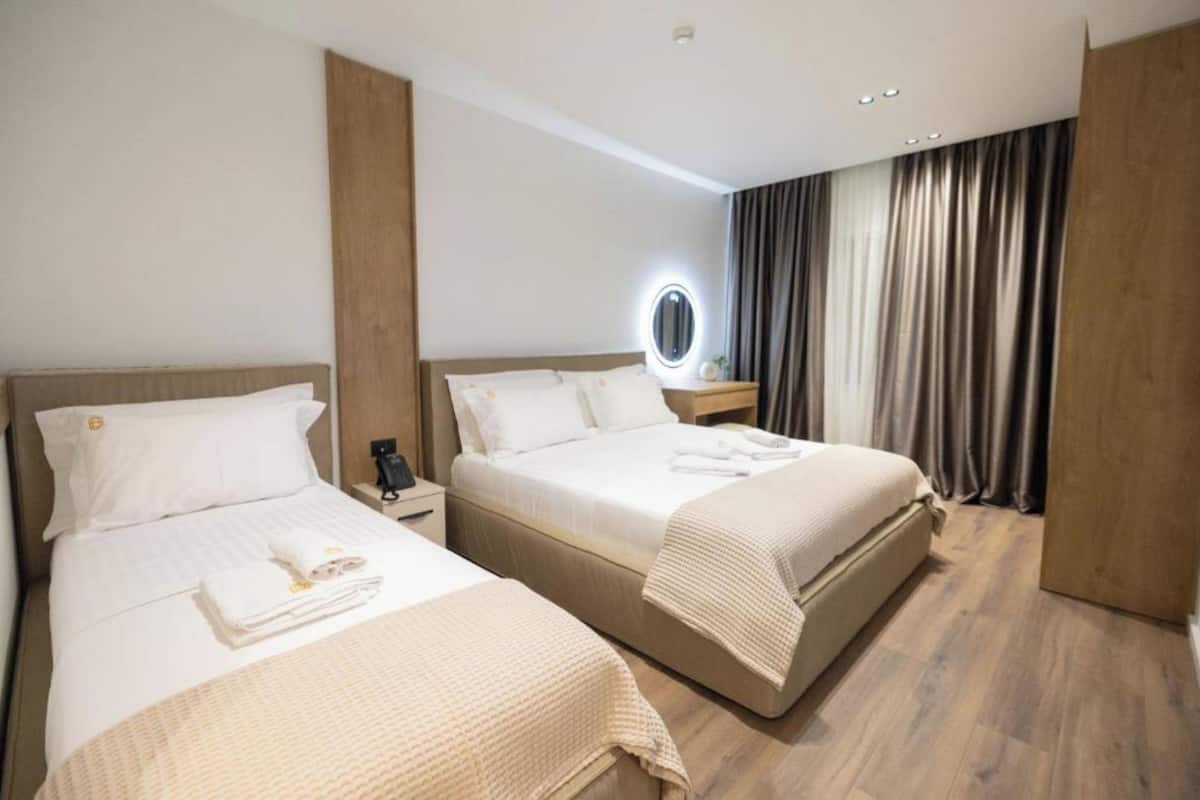 Habitación de hotel con dos camas, una mesita de noche con lámpara, un espejo y un ventanal con cortinas.