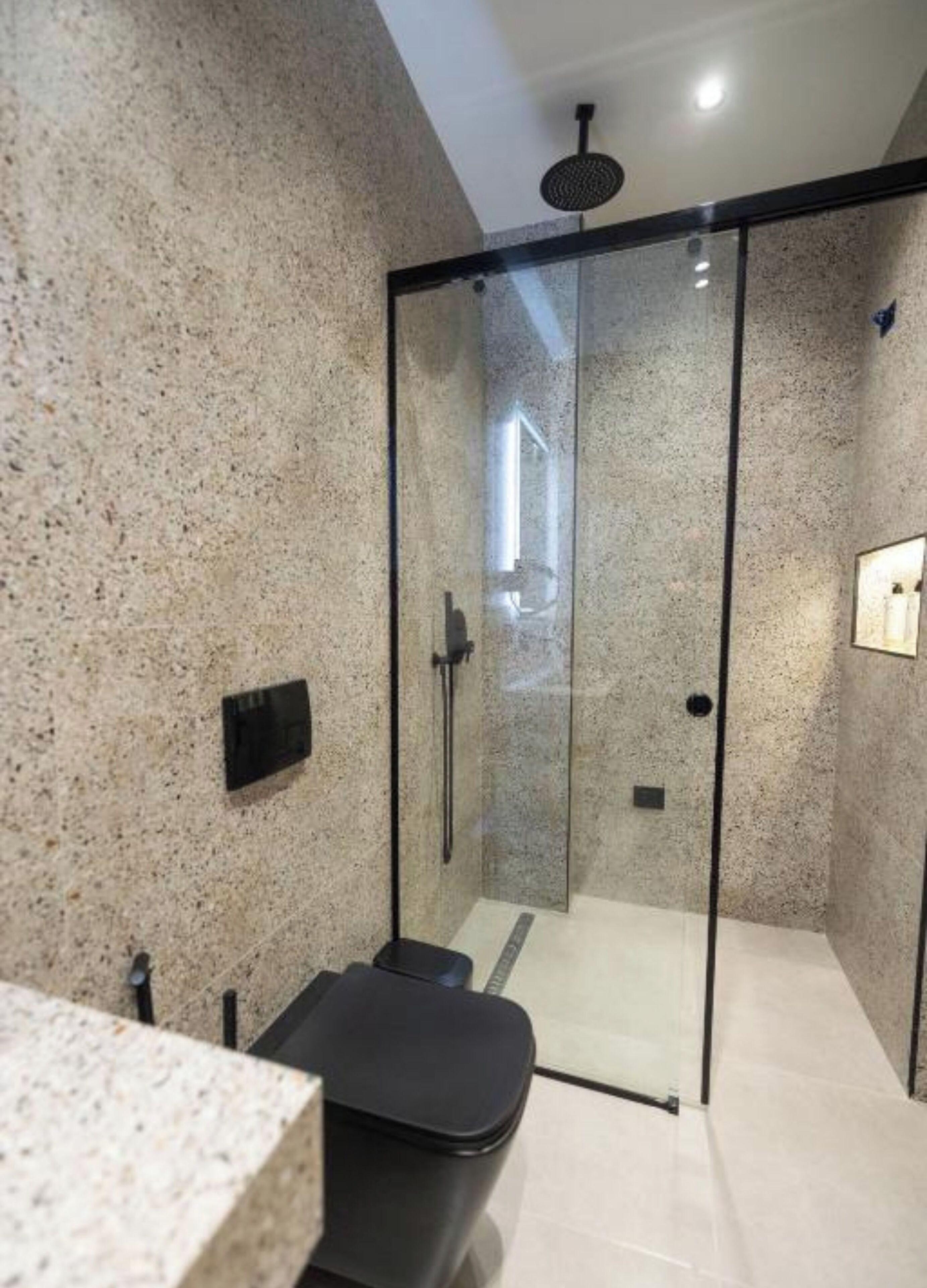 Un baño moderno con ducha envuelta en vidrio, paredes de granito y un banco negro.