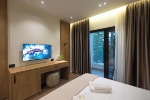 Une chambre d’hôtel moderne, dotée d’un grand téléviseur à écran plat fixé sur un meuble en bois, d’un lit avec des serviettes soigneusement pliées, d’un petit pouf rond et d’une porte-fenêtre coulissante donnant sur un balcon.