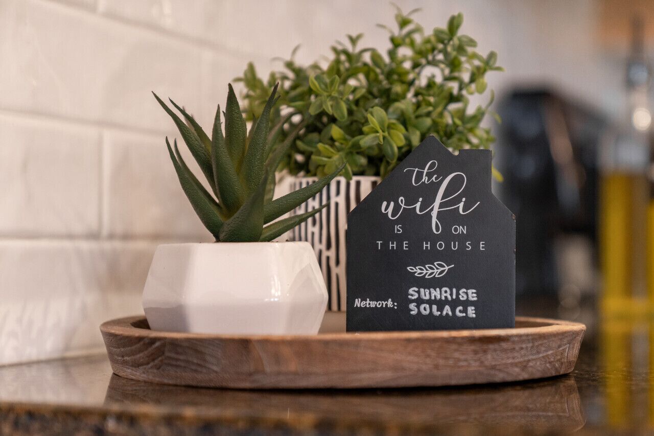 A house-shaped sign with text on a wooden tray, a white pot with a succulent plant, and a green plant in the background.