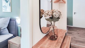 A room with a bed, a wooden table with a vase of flowers, a mirror, and a coat rack. - Garden View 1 - Design-Apartment, Küche, Garten - Sauerlanda (Schalksmühle)