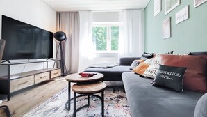 A modern living room with a flat-screen TV, a sofa with patterned cushions, a small wooden table, and a floor lamp. - Garden View 1 - Design-Apartment, Küche, Garten - Sauerlanda (Schalksmühle)