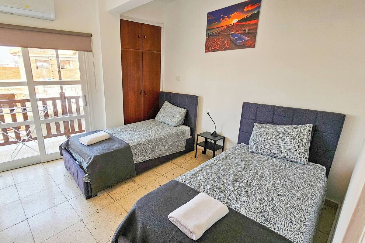 A room with two beds, a wooden wardrobe, a painting of a sunset, and a balcony with a view of buildings.