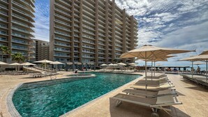 A swimming pool with lounge chairs and umbrellas, surrounded by a high-rise hotel building. - Has it all Beachfront Retreat With Stunning Views (Puerto Peñasco)