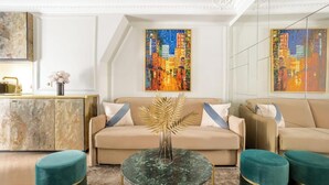 A modern living room with a beige sofa, a glass coffee table, and teal ottomans. There are two large paintings on the walls and a gold-toned cabinet.