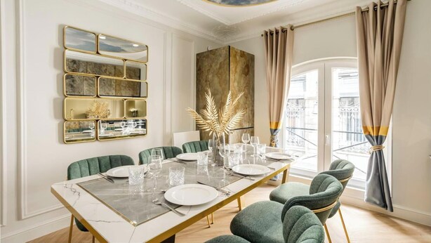 A dining room with a marble table, green upholstered chairs, and a gold-framed mirror.