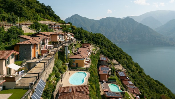 A hillside residential area with terraced houses, a swimming pool, and a view of a lake and mountains.