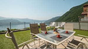 An outdoor dining area with a glass table and chairs, overlooking a lake and mountains.