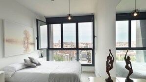 A modern bedroom with a large bed, a view of the city through large windows, and a sculpture on the nightstand. - Unik Vacation The Tower (Tarragona)