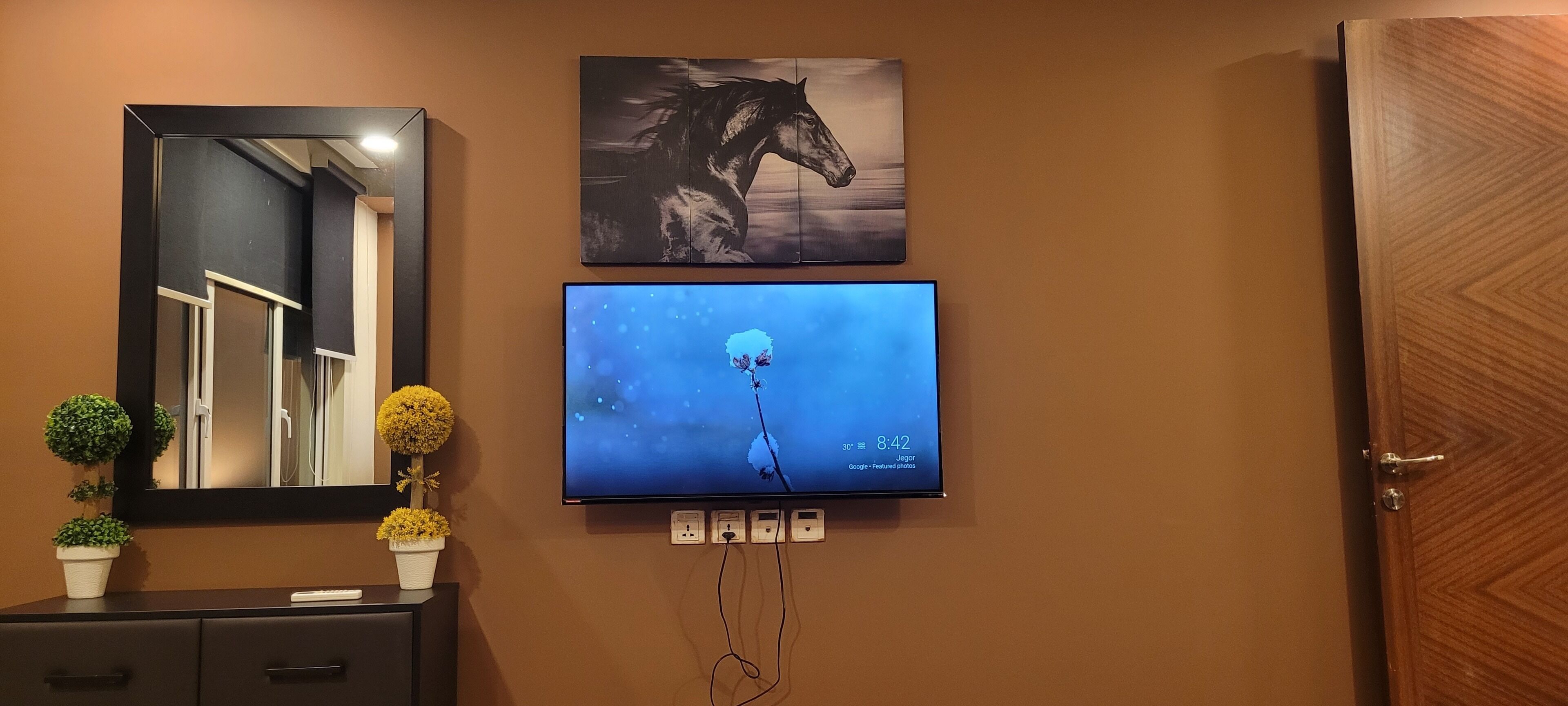 Un televisor de pared muestra una pantalla azul con una flor y un reloj, un espejo y una puerta.