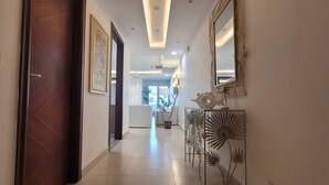Un pasillo con una puerta de madera, un interruptor de luz en la pared, un cuadro enmarcado y un atril metálico decorativo con un espejo.