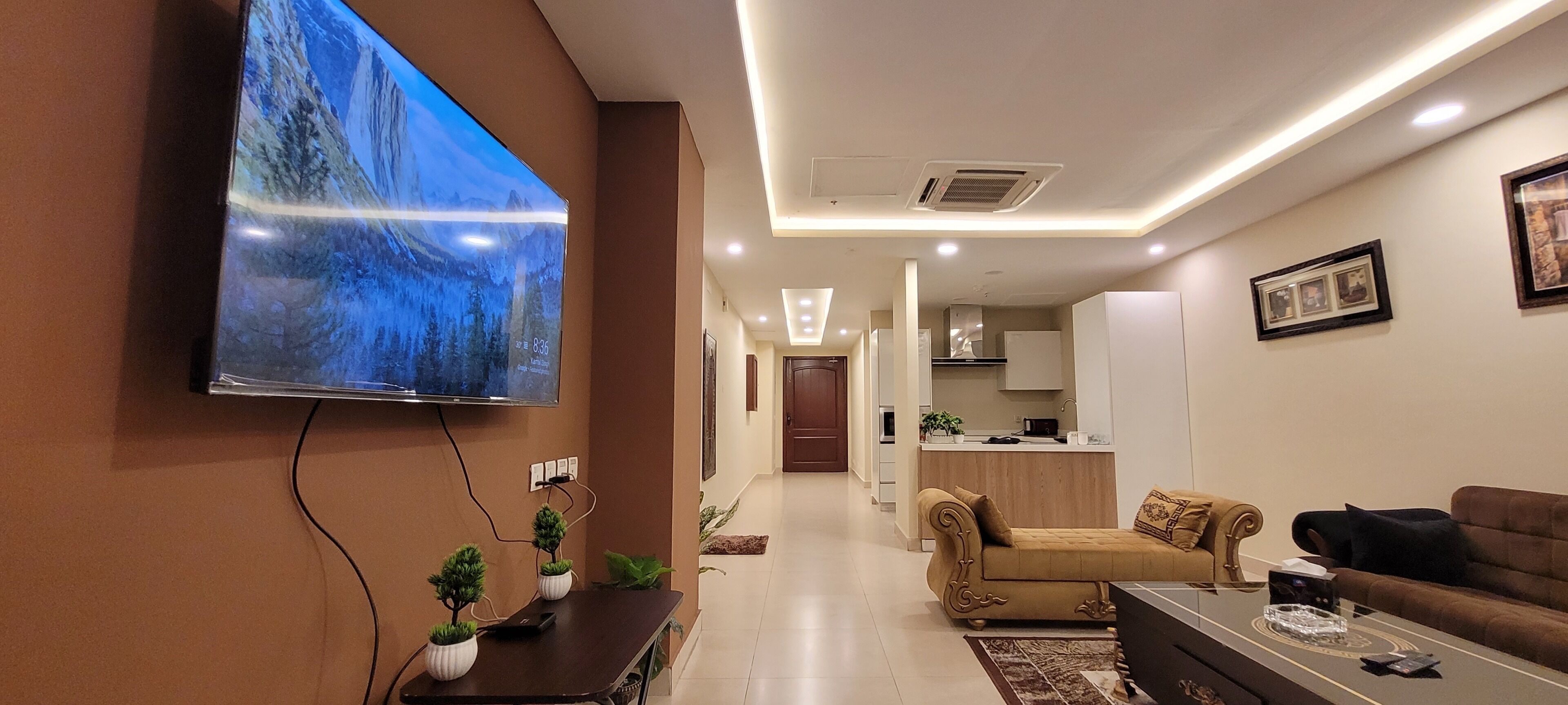 A modern living room with a flat-screen TV, a sofa, a coffee table, and a kitchen area in the background.