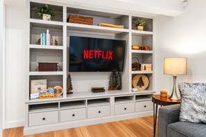 A modern living room with a flat-screen TV mounted on a white entertainment unit, surrounded by shelves with decorative items and books.