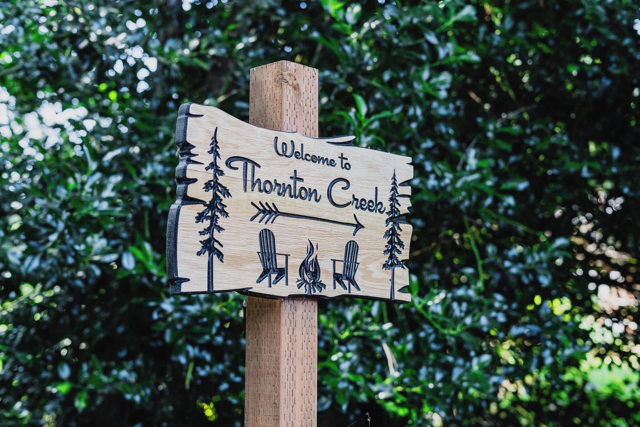 Un letrero de bienvenida de madera para Thornton Creek, con un diseño rústico que incluye árboles e ilustración de una fogata.