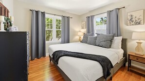 A bedroom with a bed, curtains, a bedside table, and a window. - Charming Historic Cottage • Cozy Family Getaway! (Greeley)