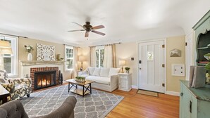 A living room with a fireplace, a ceiling fan, a sofa, a coffee table, and a patterned rug. - Charming Historic Cottage • Cozy Family Getaway! (Greeley)