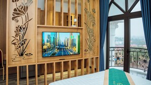 A hotel room with a large TV mounted on a wooden panel with floral carvings, a balcony with a view of the city, and a bed with a green runner. - Gala Hotel & Apartment (Hai Phong)