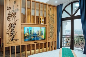 Et hotelværelse med stort TV monteret på en træpanel med blomstermotiver, en balkon med udsigt over byen og en seng med grøn sengetæppe.
