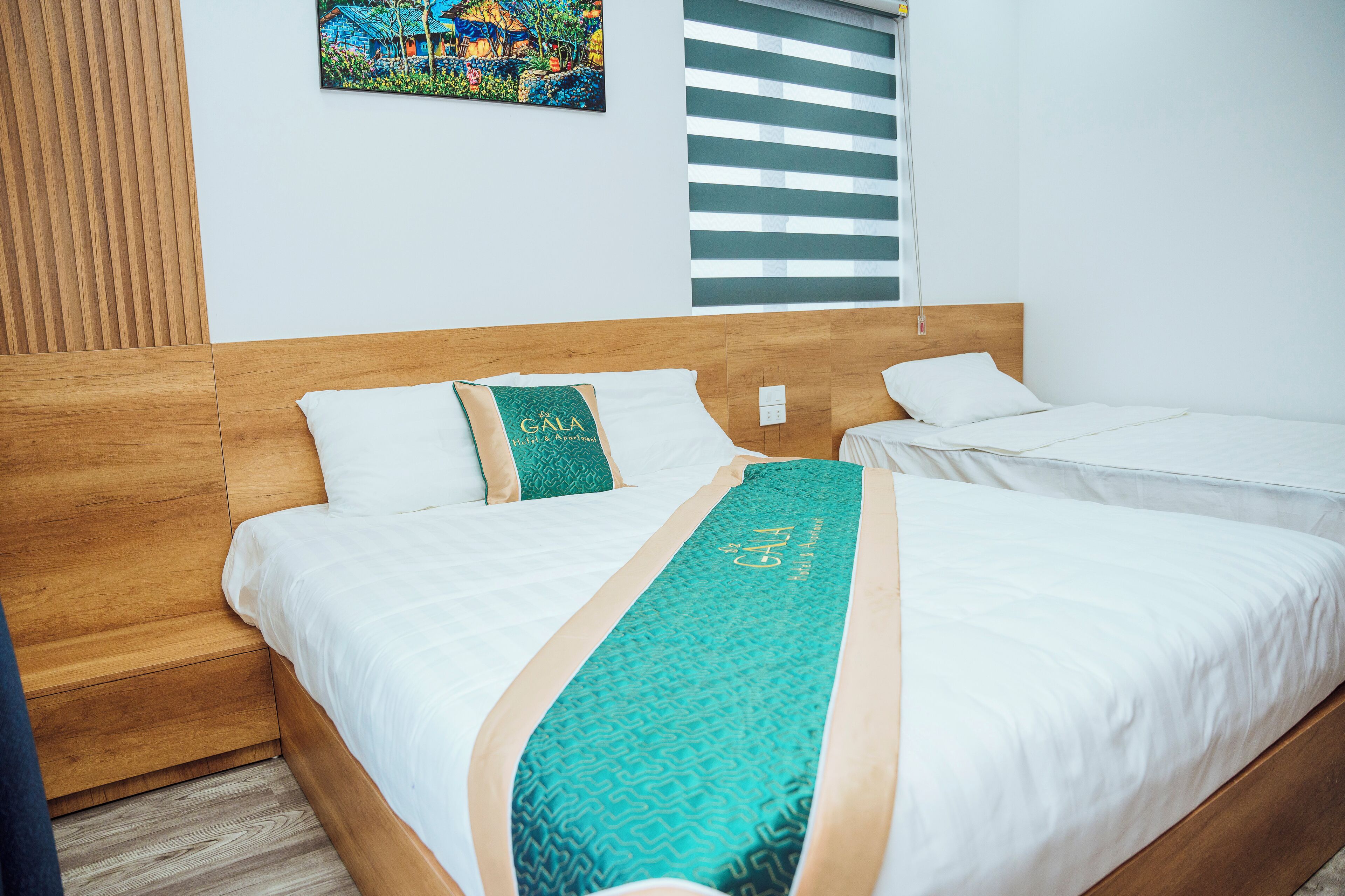 Ein Hotelzimmer mit zwei Betten, einem Holzkopfstück, einem türkisfarbenen Bettüberwurf und einem farbenfrohen Gemälde an der Wand.