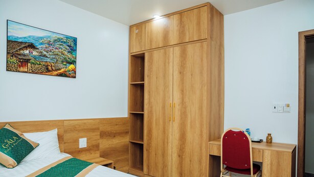 A hotel room with a bed, a wooden wardrobe, a desk with a chair, and a painting on the wall. - Gala Hotel & Apartment (Hai Phong)
