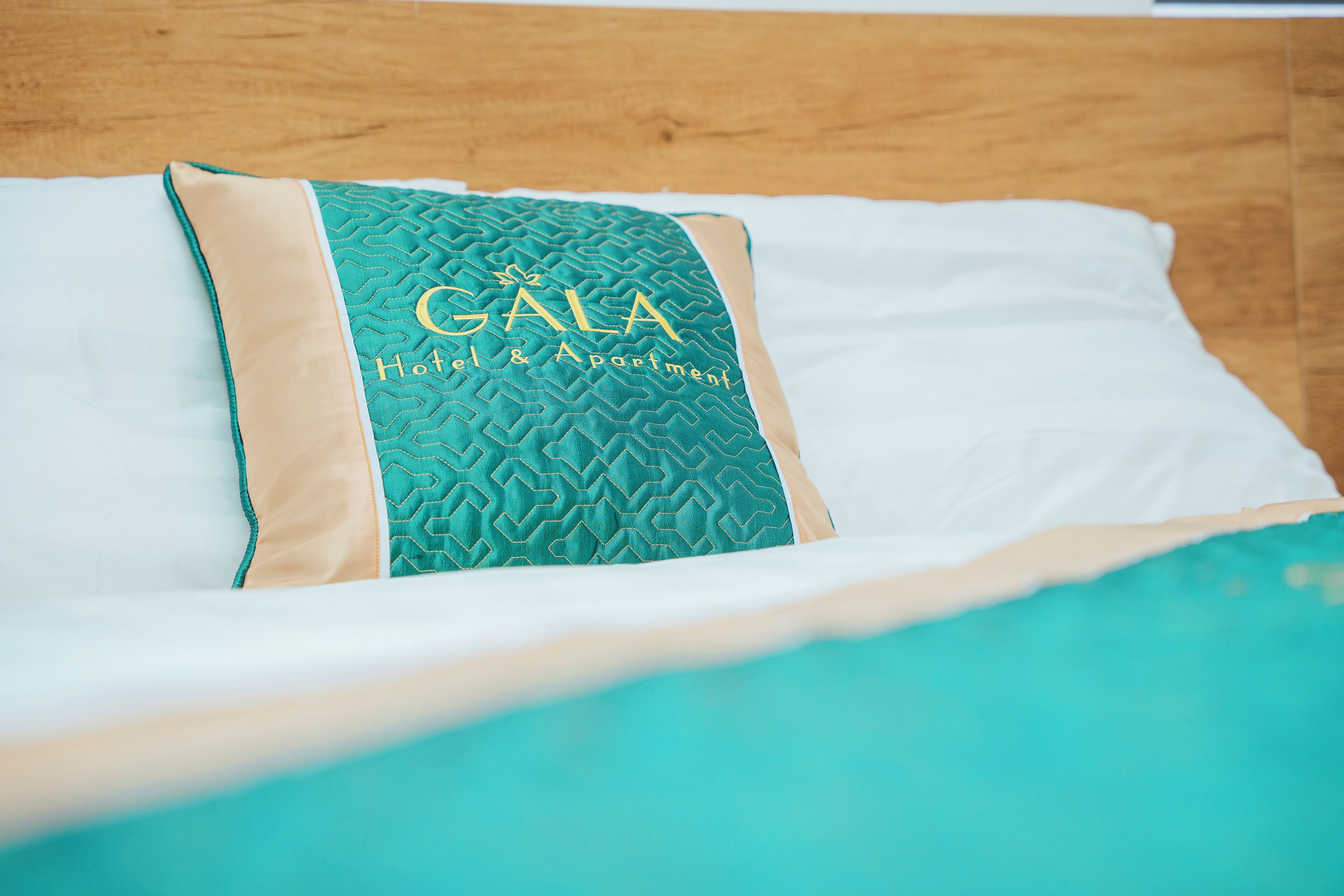 A bed with a teal and beige pillow featuring the text "GALA Hotel & Apartment".
