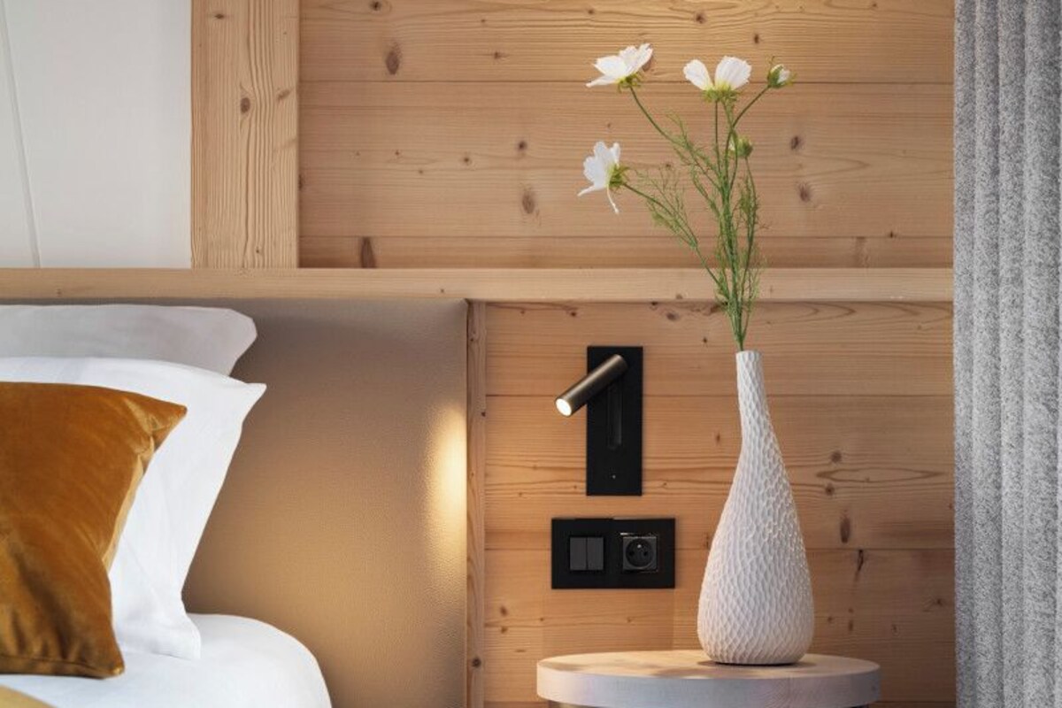 A modern bedroom with a wooden headboard, a bed with white and mustard bedding, a round side table with a white vase, and a wall-mounted light fixture.