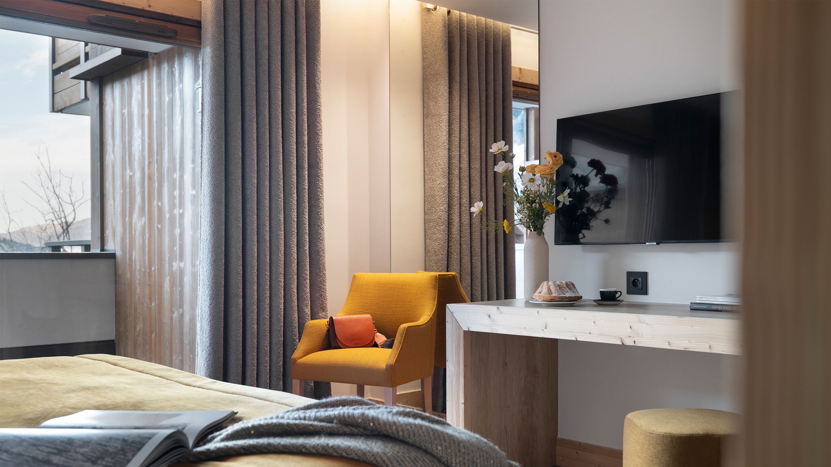A modern hotel room with a large window, a comfortable yellow armchair, a bedside table with a vase of flowers, and a flat-screen TV mounted on the wall.