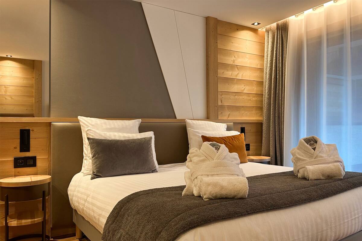 A modern hotel room with a large bed, wooden headboard, and a view of the city through sheer curtains.