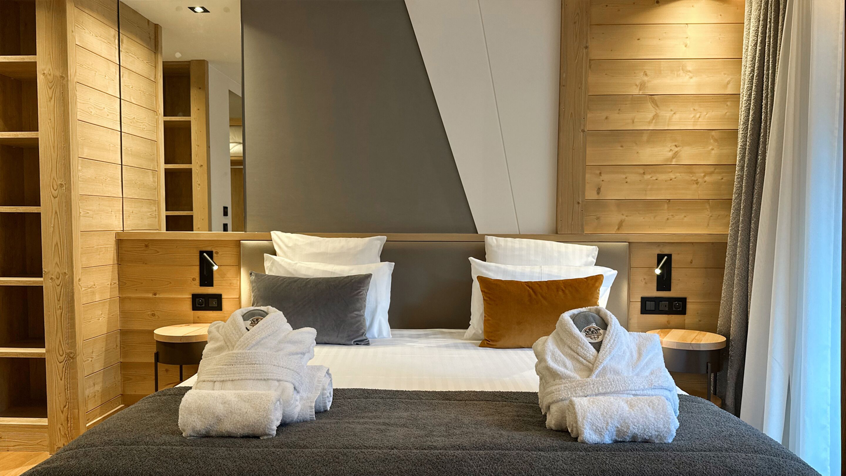 A modern hotel room with a large bed, two bedside tables, a wooden wardrobe, and a mirror.