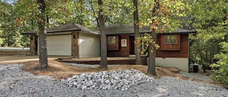A cabin with a garage, surrounded by trees and a gravel driveway.