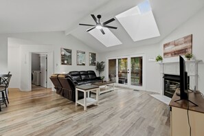 A modern living room with a black leather sofa, a coffee table, and a TV. There is a fireplace and a ceiling fan. - Bike In/Bike Out Home in Bella Vista (Bella Vista)