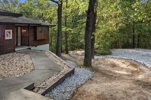A wooden cabin with a concrete pathway leading to the entrance, surrounded by a forested area with gravel paths.