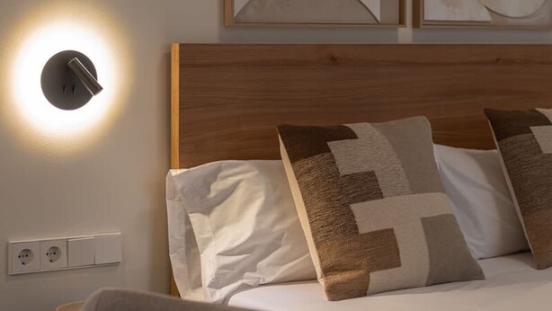 A neatly made bed with patterned pillows, a bedside table, and a wall-mounted light fixture. - Sant Pau Patio 2BR Near Avinguda Gaud With Patio (Barcelona)
