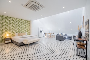 Une chambre d’hôtel moderne avec un sol à motifs, un lit doté d’une tête de lit blanche, un canapé gris, un coin repas et un mur recouvert d’un papier peint à motifs de feuilles.