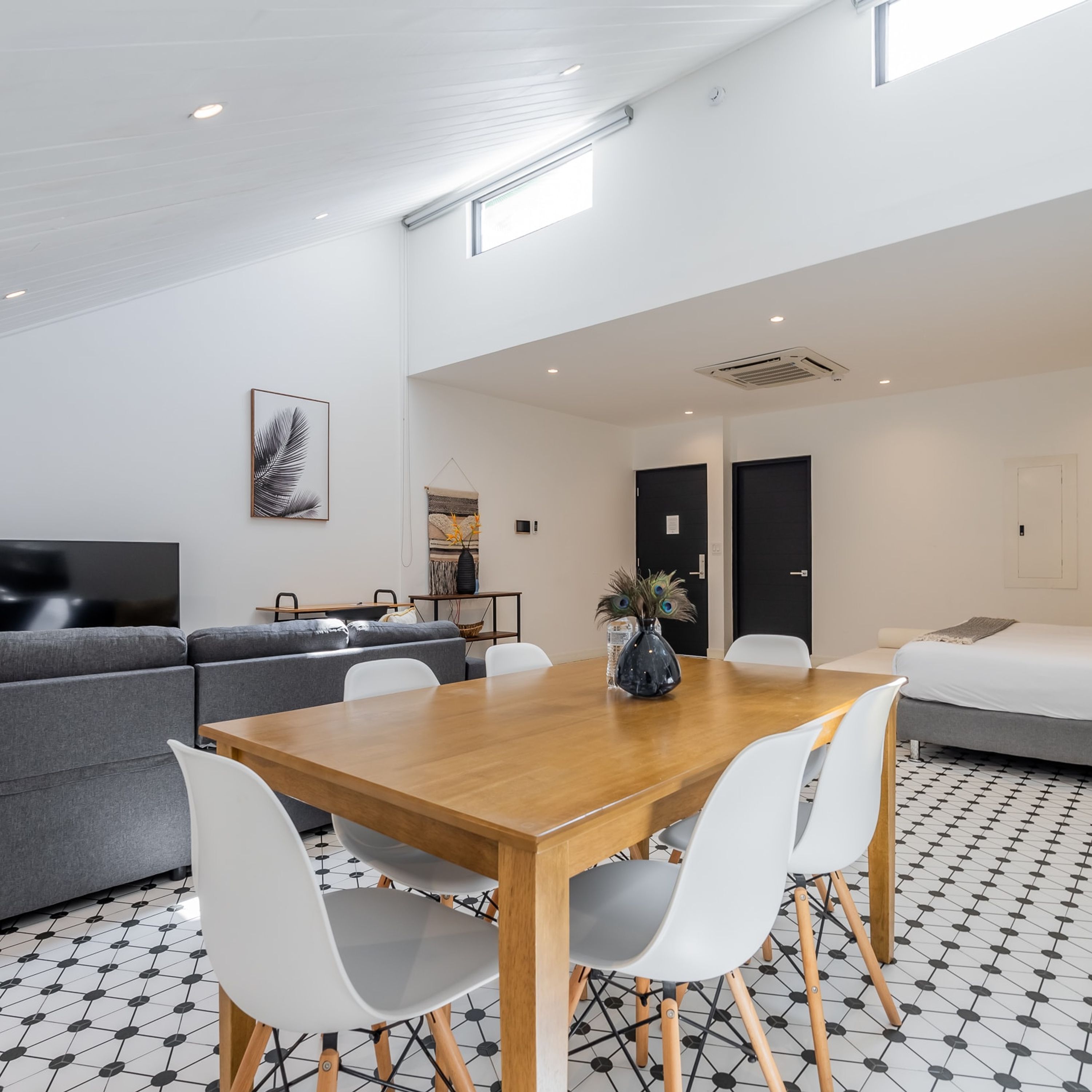 A modern, well-lit room with a wooden dining table, white chairs, a grey sofa, and a bed.