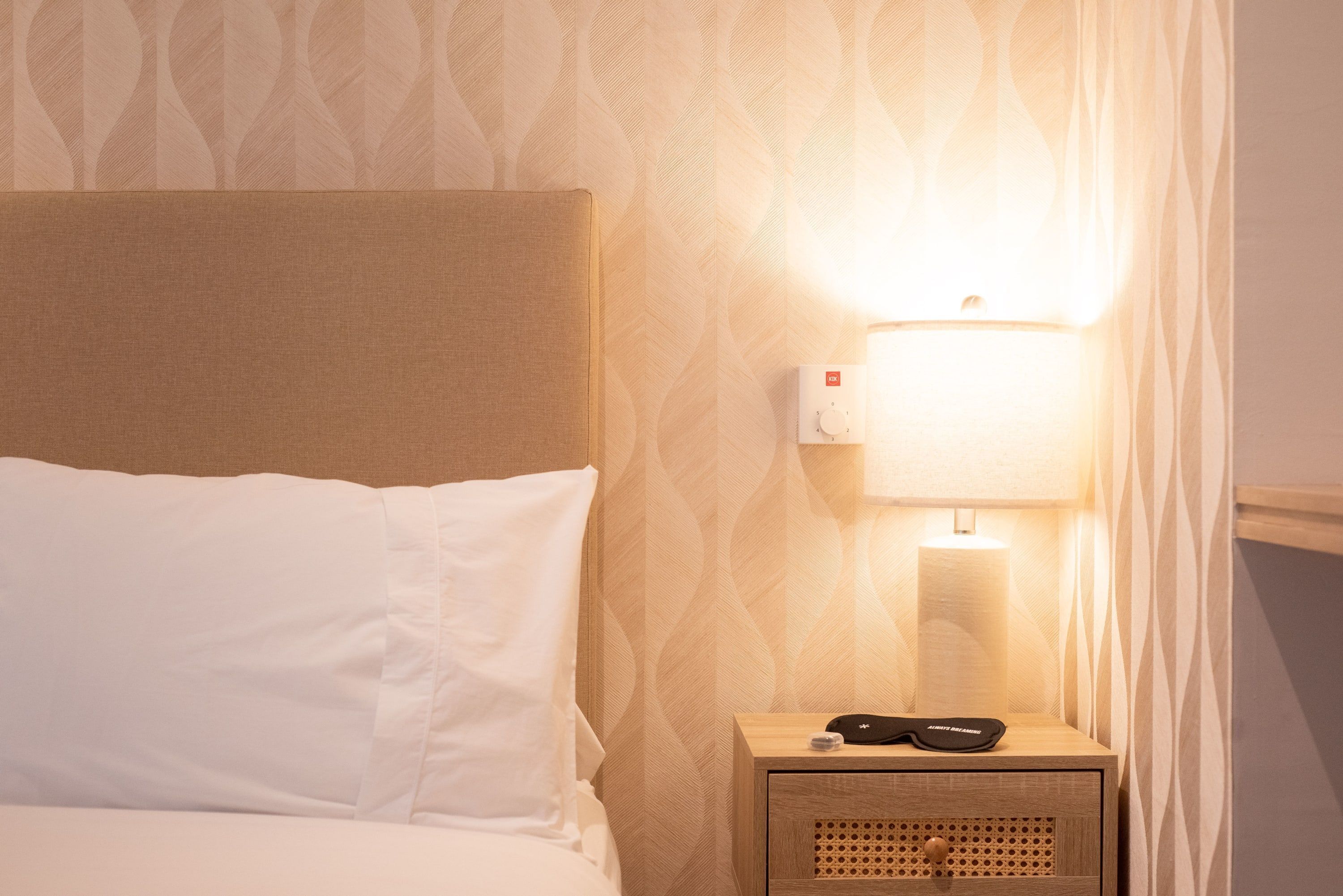 Een net opgemaakt bed met witte lakens, een houten nachtkastje met een lamp, en een kamer met een patroon op de muur.