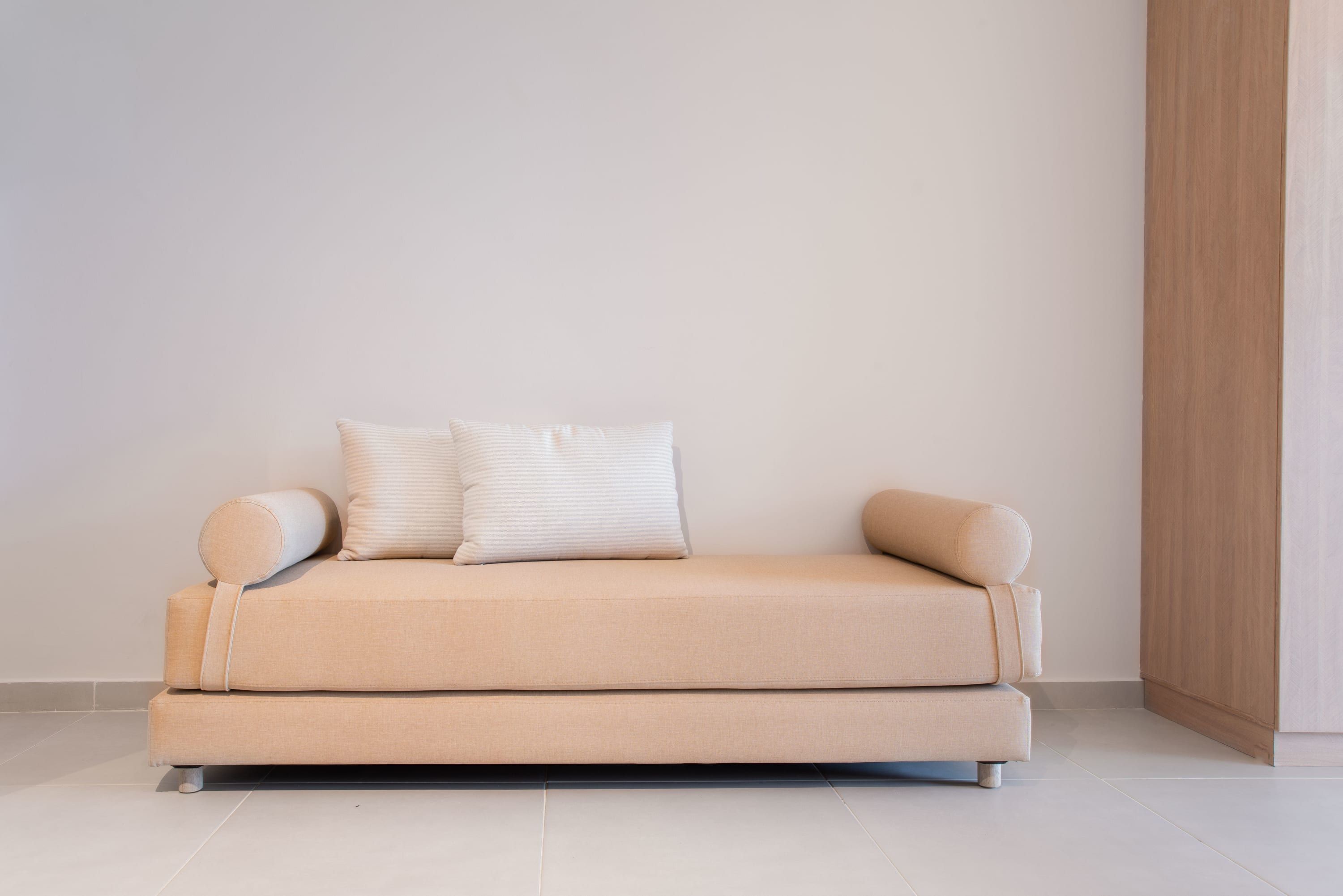 Een minimalistische beige bank met twee kussens en een passende voetenbankje in een hoek van een kamer met effen witte wanden en een houten vloer.