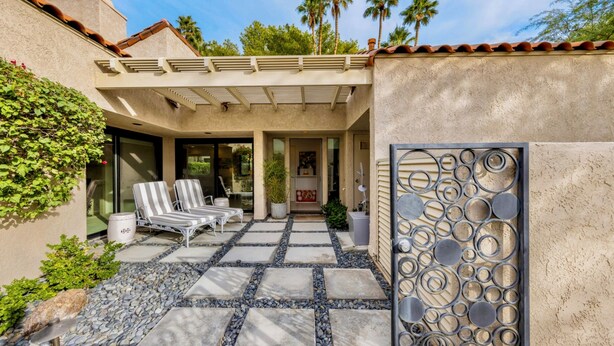 A covered patio with two striped lounge chairs, a stone pathway, and a decorative metal gate.