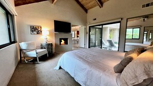 A bedroom with a large bed, a chair, a table with a lamp, a fireplace, and a sliding glass door leading to a balcony with lounge chairs.