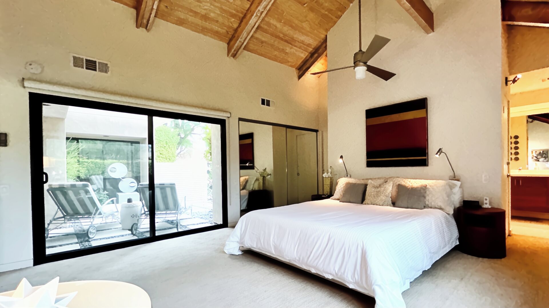 A bedroom with a large bed, a ceiling fan, a sliding glass door leading to a patio with outdoor furniture.