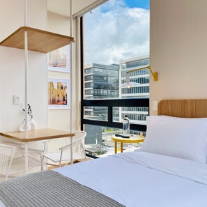 A hotel room with a bed, a desk, a chair, a small table with a bottle and remote, and a view of modern buildings. - w Magnificent Loft With Perfect View (Bogotá)