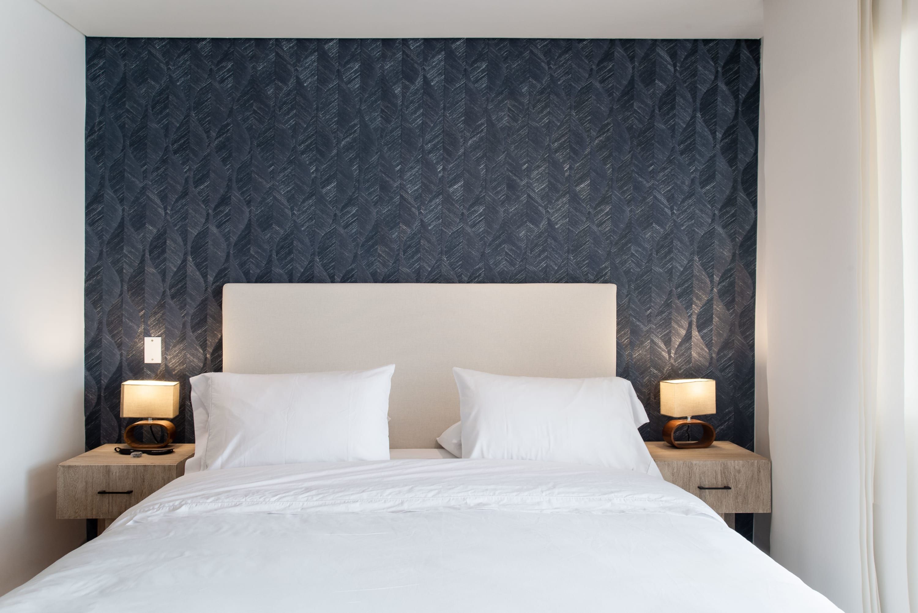 Un lit bien fait, avec du linge de lit blanc, deux tables de chevet avec des lampes et un papier peint à motifs sur le mur derrière le lit.